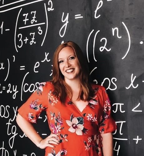Holly in an orange-flowered dress, standing in front of a black chalkboard with white math equations on it